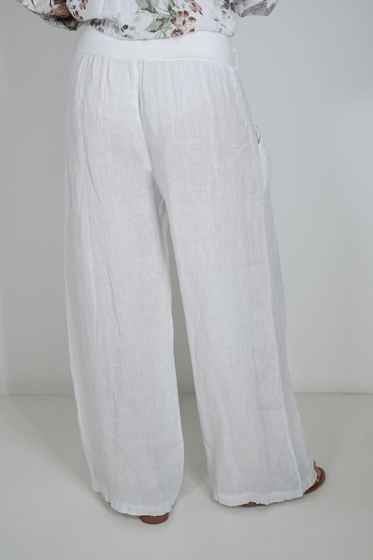 White wide-leg linen pants shown from the back on a model, paired with a floral print top, against a plain white studio background.
