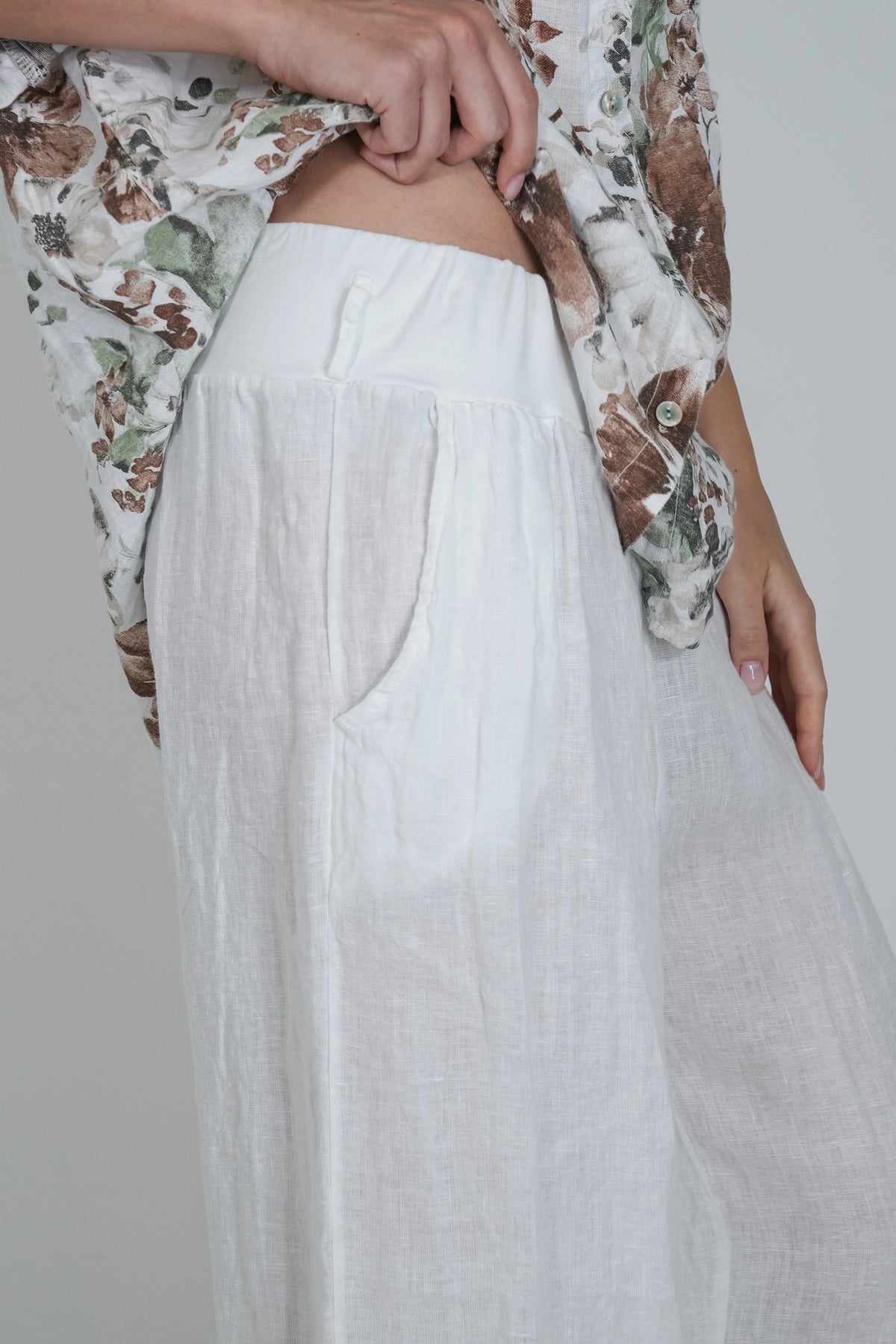 White linen wide-leg trousers with side pockets, shown on a model wearing a floral shirt against a light gray background.