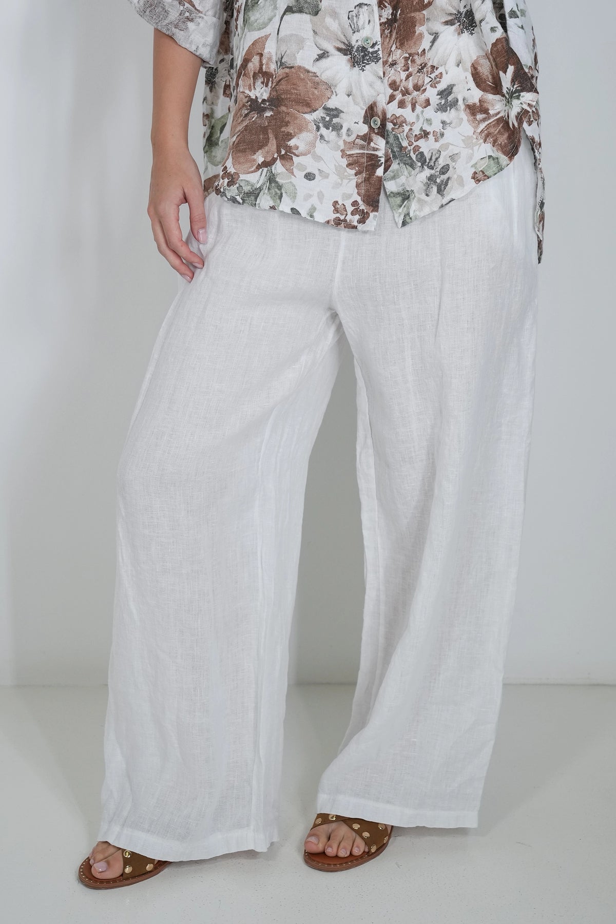 A person wearing loose white wide-leg linen pants, paired with a brown and white floral print top and brown sandals, standing against a plain white studio background.