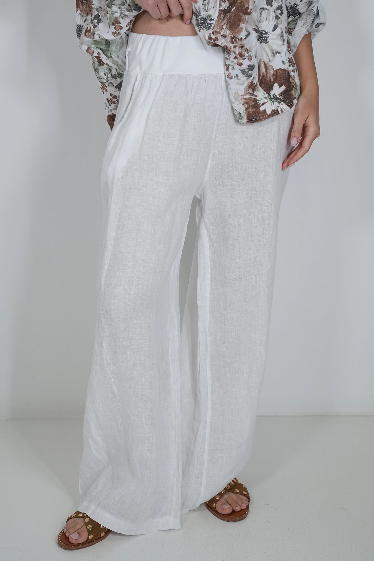 White wide-leg linen pants worn by a model, styled with a brown and white floral shirt and brown slide sandals against a plain white studio background.
