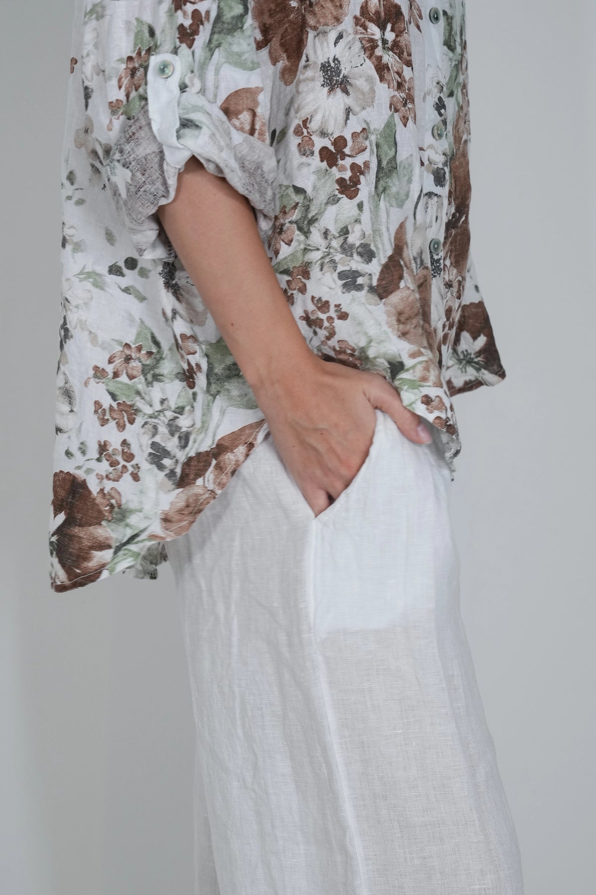 A person wearing a loose white blouse with a brown and green floral print and white wide-leg pants, standing against a plain light gray background.