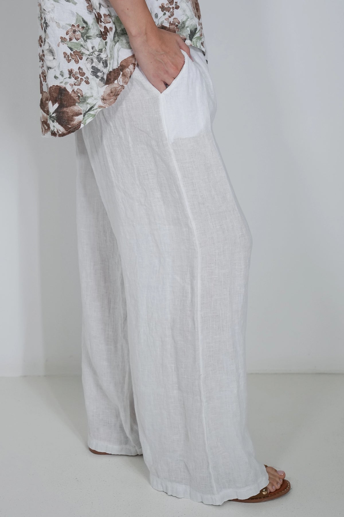 A person wearing loose, wide-leg white linen pants with a side pocket, paired with a floral top and brown sandals, standing against a plain white wall.