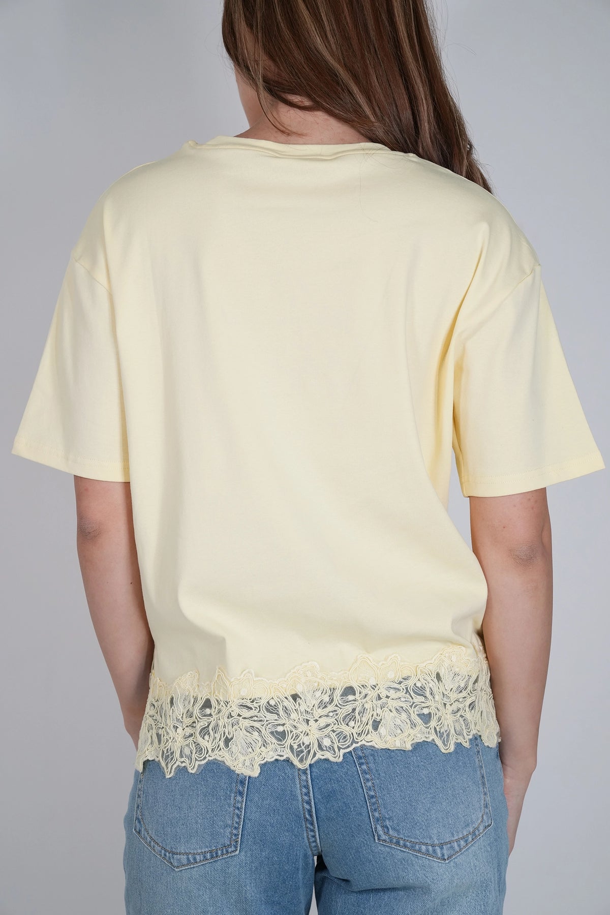 A person shown from the back wearing a pale yellow short-sleeve T-shirt with a decorative lace hem, paired with blue jeans, against a plain light gray background.