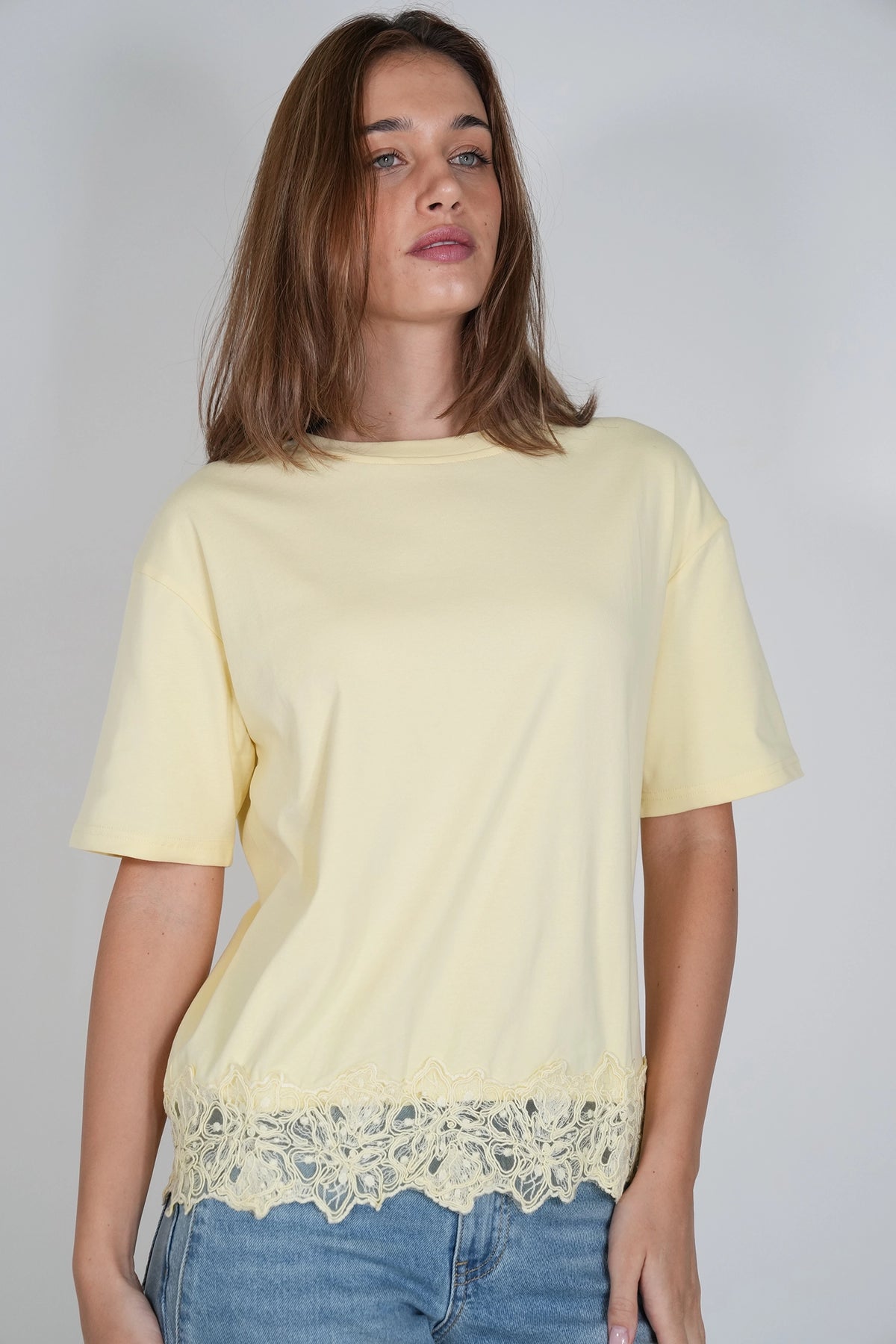 A woman wearing a pale yellow short-sleeve t-shirt with a decorative cream lace hem, paired with blue jeans, against a plain light gray background.
