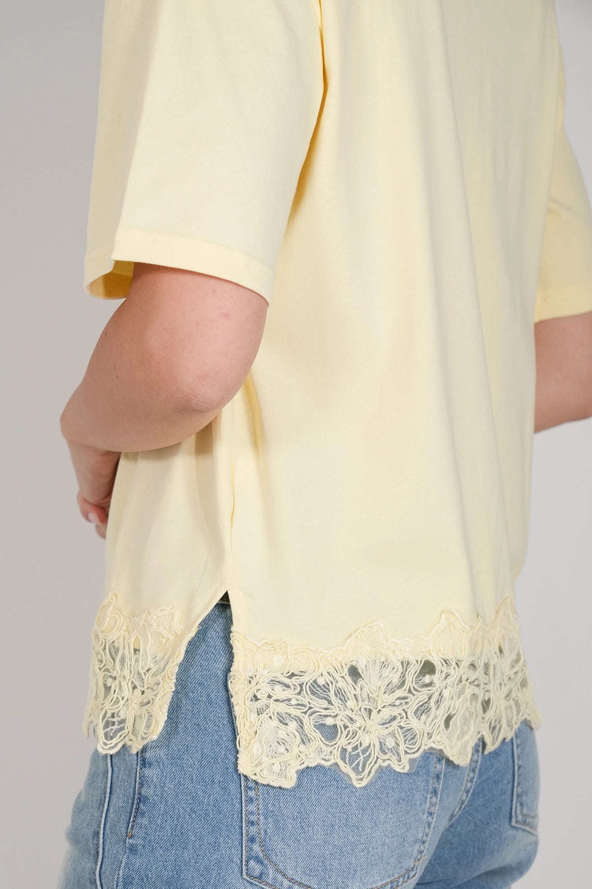 A close-up of a person wearing a pale yellow short-sleeve top with a floral lace hem, paired with light blue jeans, against a neutral gray background.