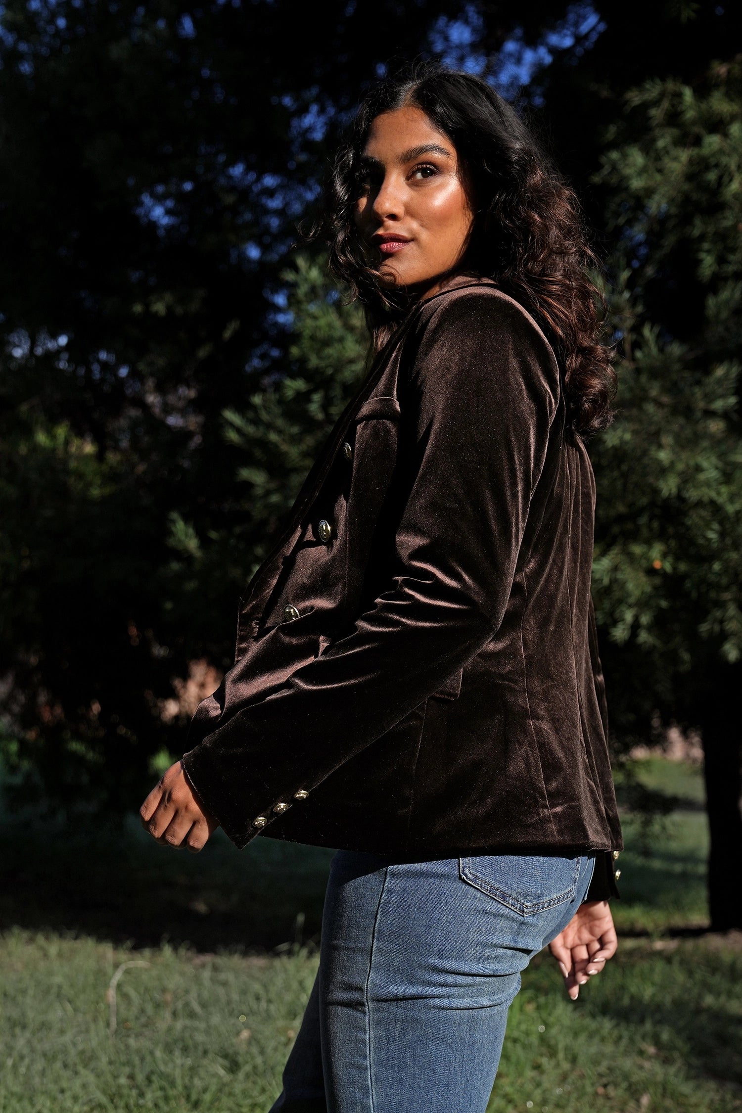 Woman wearing a brown jacket and blue jeans standing outdoors with trees in the background