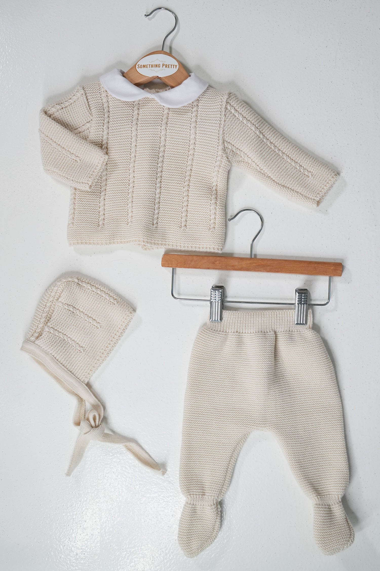 Baby sweater, hat, and pants set on hangers against a white background