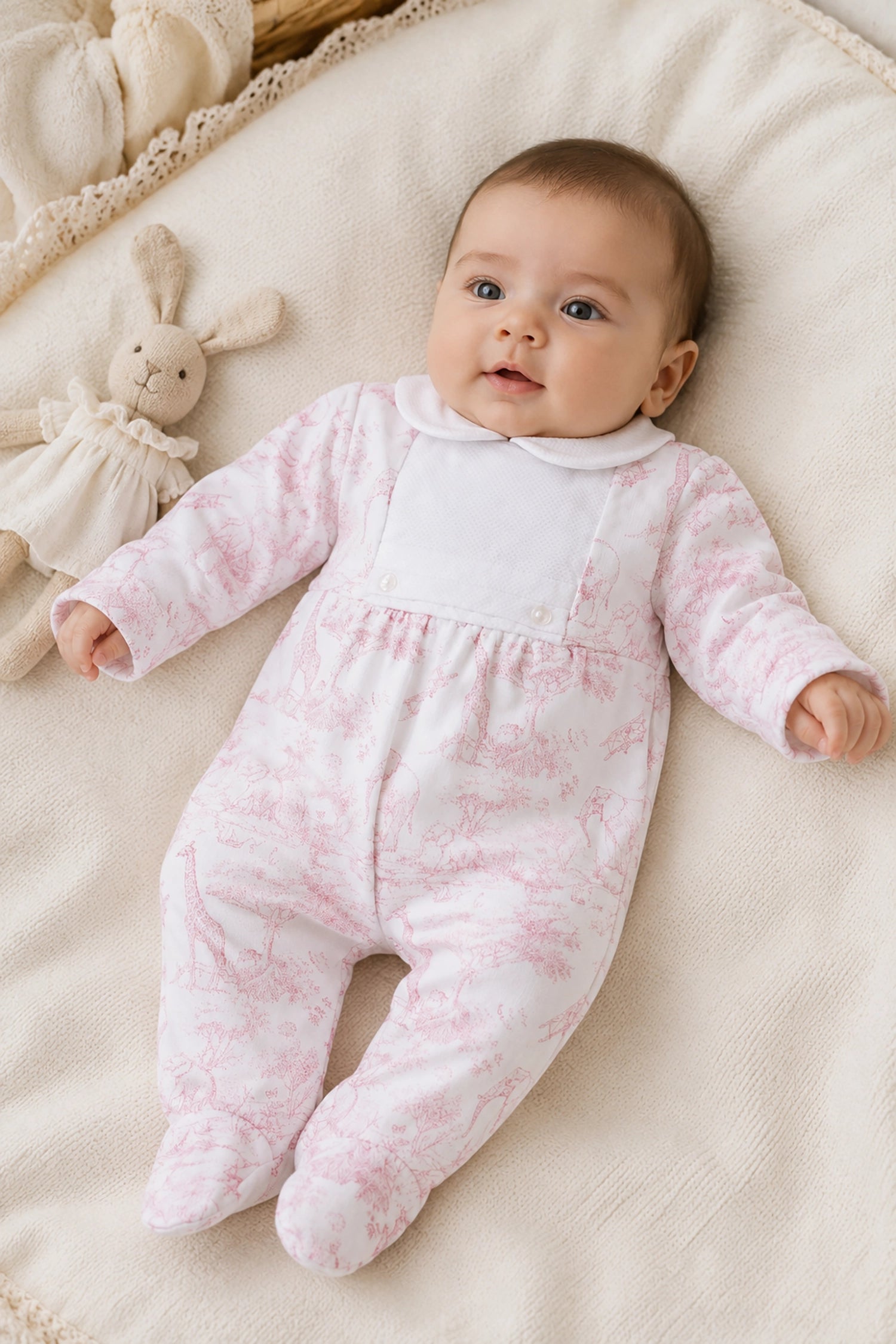 Baby in a pink and white patterned onesie lying on a soft surface with a plush toy.