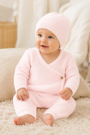 Baby wearing a pink knitted outfit and hat sitting on a soft surface.